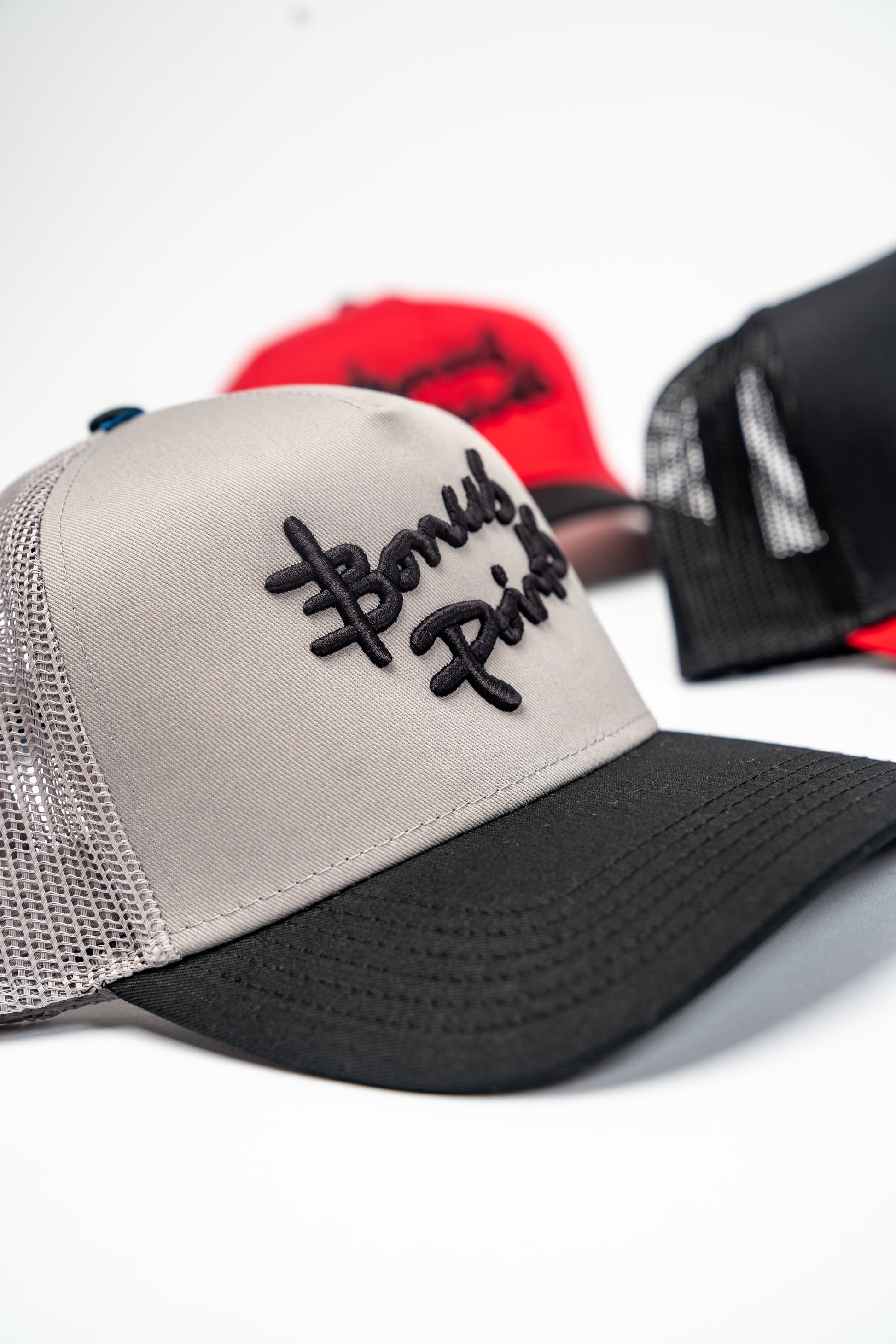 Side Close up Shot of Gray and Black “Extra Credit” Trucker Hat amongst a few other trucker hats  in the background 